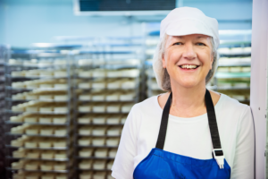 International Women's Day. Stacey Hedges, maker of Tunworth and Winslade, is standing to the right of the frame wearing a white shirt and hat, and a blue apron. She is smiling to the camera. Behind her are racks of maturing cheeses.